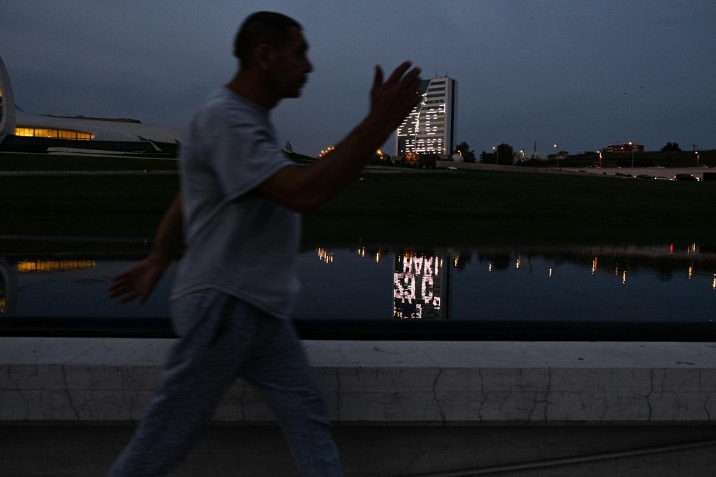 A man in motion walking along a river, silhouetted against the evening city skyline.