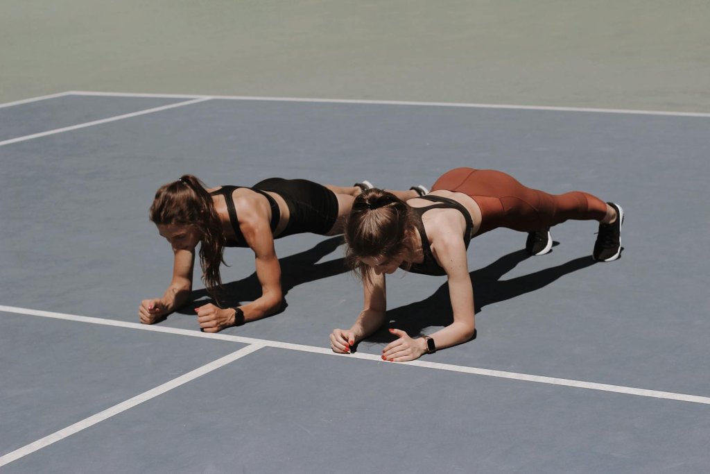 Two women in activewear doing plank exercises on an outdoor sports court.
