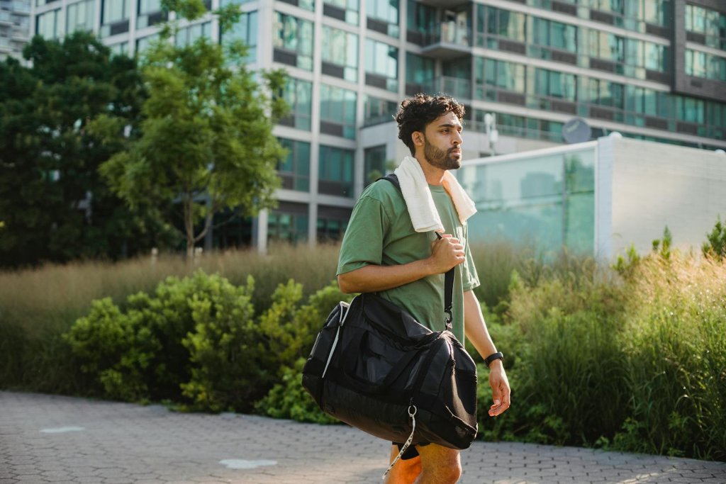 A man walks through a city park carrying a sports bag, with a towel draped over his shoulder.