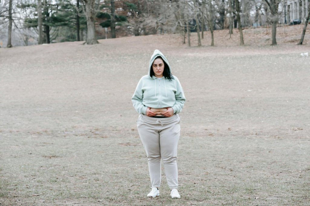Full length of unhappy young woman in activewear standing in autumn park and showing fat on waist