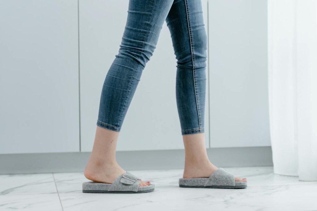Close-up of a person wearing jeans and gray slippers, walking indoors.