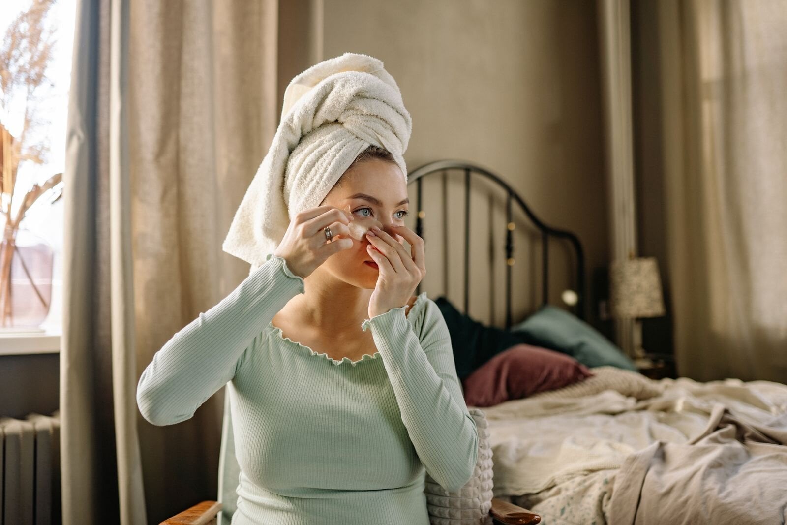 A woman applies beauty products while practicing self care in a cozy, well-lit bedroom.